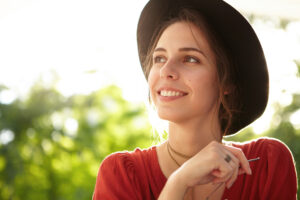 Horizontal portrait of pleased young female wearing elegant red dress and black hat standing in park admiring summer vacations. Sexy woman with smile looking aside having mystreious expression